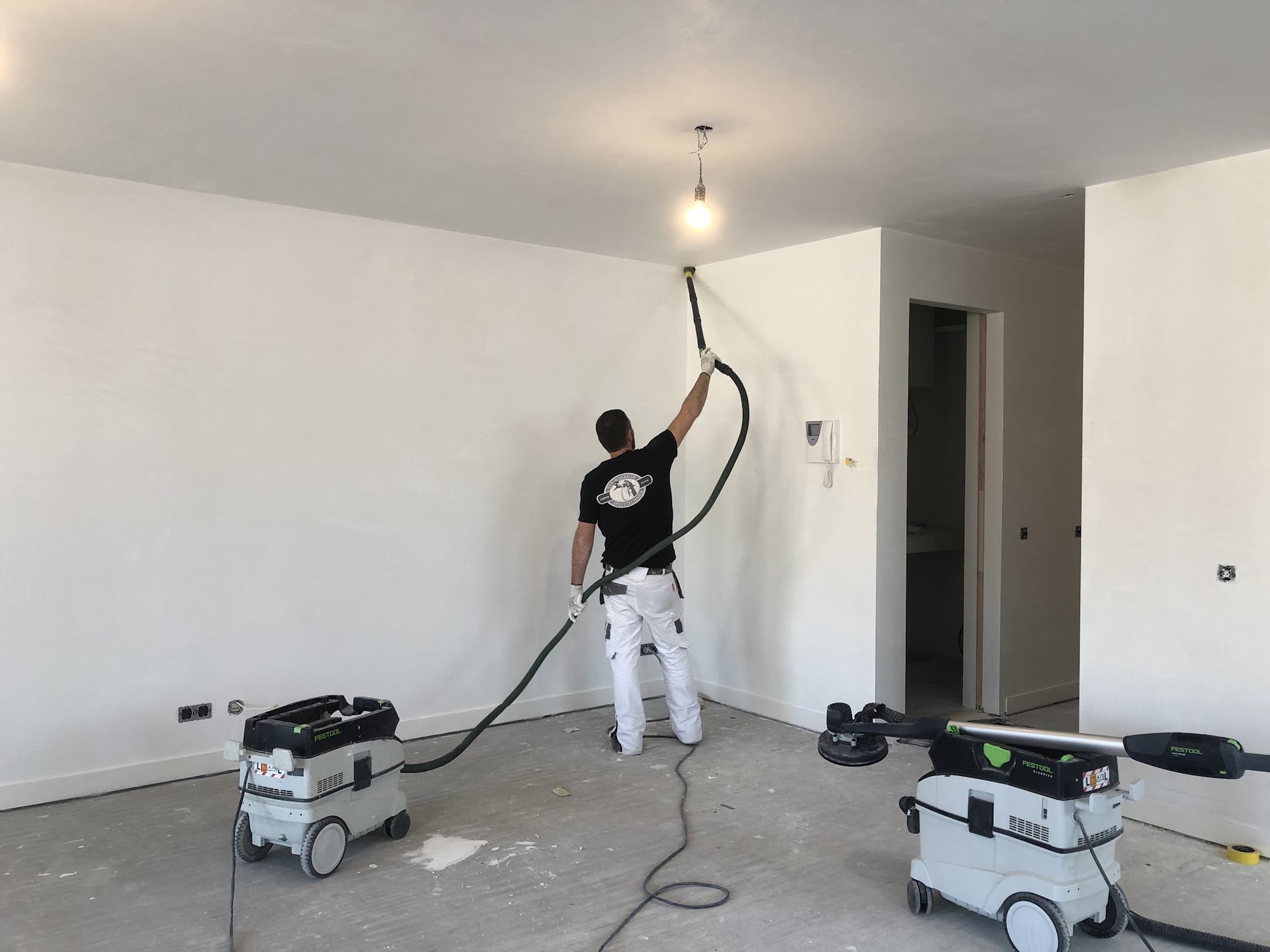 Een van de mannen schuurt een wand stofvrij met een langarmschuurmachine en twee stofzuigers